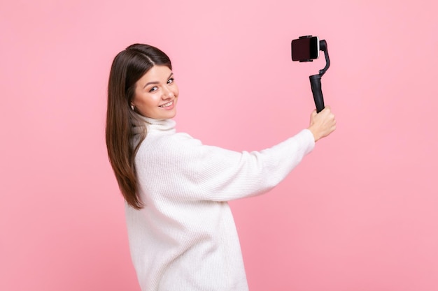 Fêmea usando steadicam e telefone celular para transmissão ao vivo olhando para a câmera com sorriso vestindo suéter branco estilo casual Tiro de estúdio interior isolado no fundo rosa