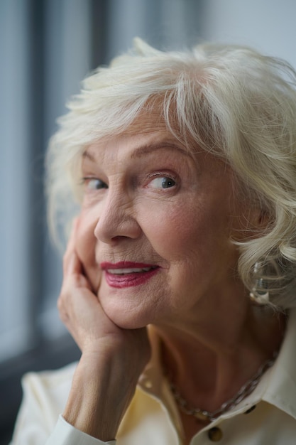 Feminidad. Cara de primer plano de una anciana hermosa de cabello gris con labios rojos tocando la mano a la cara mirando la cámara a la luz del día