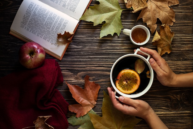 Femininas mãos segurando uma caneca de vinho quente quente. vinho quente, mel, folhas de outono, maçãs, um livro e uma blusa de lã sobre um fundo de madeira. clima aconchegante de outono. fundo de outono. configuração plana