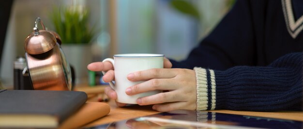 Femininas mãos segurando uma xícara de café na mesa de escritório em casa de madeira com agenda livros, suprimentos e decorações