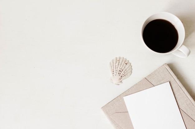 Feminine Briefpapier-Desktop-Mockup-Szene Leere Grußkarte Bastelumschlag Tasse Kaffee Muschel und alte Bücher auf weißem Tischhintergrund Flache Ansicht von oben Sommer Herbst rustikale Komposition