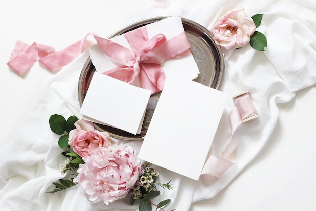 Feminine Hochzeit Geburtstag Mockup Szene Blankopapier Grußkarten Umschlag Eukalyptus rosa Rosen Pfingstrose Blumen und Bänder auf weißem Tischhintergrund Leichtes Schattenspiel Flache Ansicht von oben