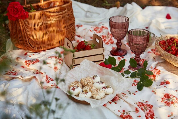 Feminines rosa Sommerpicknick mit Tischdecke, Marshmallows, Erdbeeren, Kirscherbsen und Weingläsern zwischen rosa Rosen im Park. Romantische Abendreise und Wertschätzung des Augenblicks