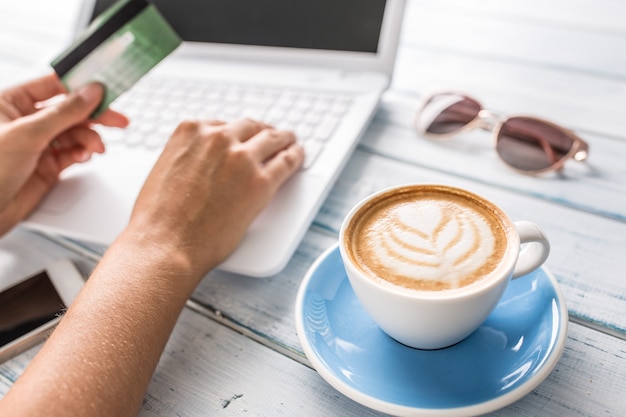 Feminino mão segurando o cartão de crédito usando laptop e café art em primeiro plano.