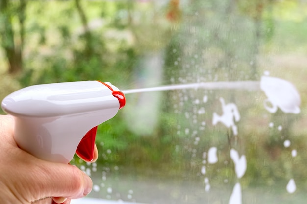 Fensterscheibe mit Sprühreiniger reinigen. Vor dem Fenster verschwommene Straße, Grün. Konzept für Hausreinigungsdienste