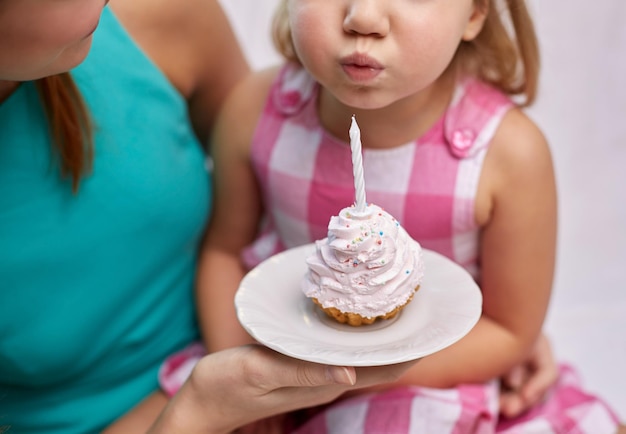 Feriado, celebração, família, aniversário e conceito de pessoas - mãe feliz segurando cupcake e garotinha soprando vela