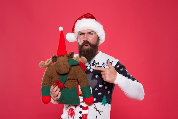 Foto feriado de inverno. conceito de preparação de natal. compre presentes. venda e compras de inverno. festa de inverno. brinquedo de rena. homem com chapéu de papai noel segurar fundo vermelho de presente de natal. suéter de inverno hipster de rosto alegre.