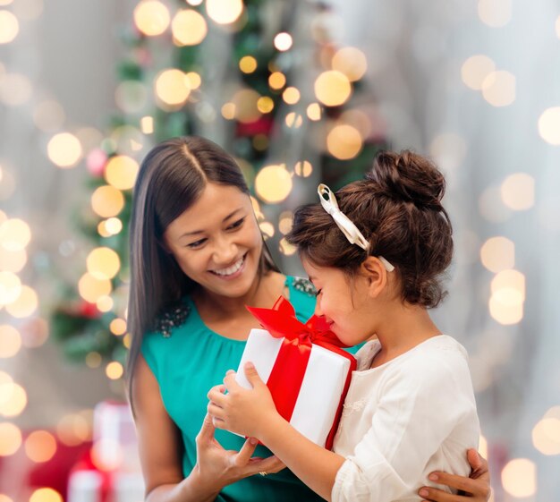 feriados, celebração, família e conceito de pessoas - feliz mãe e garotinha com caixa de presente sobre a sala de estar e o fundo da árvore de natal