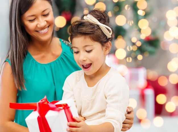 feriados, celebração, família e conceito de pessoas - feliz mãe e garotinha com caixa de presente sobre a sala de estar e o fundo da árvore de natal