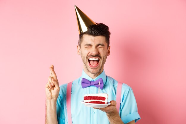 Feriados e conceito de celebração. Cara animado, comemorando o aniversário e fazendo desejo, cruzar os dedos para dar boa sorte enquanto segura o bolo de aniversário com vela, fundo rosa.