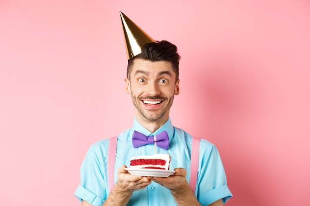 Feriados e conceito de celebração. Jovem alegre comemorando aniversário com chapéu de festa, segurando um bolo de aniversário com vela e fazendo desejo, sorrindo feliz para a câmera, fundo rosa.
