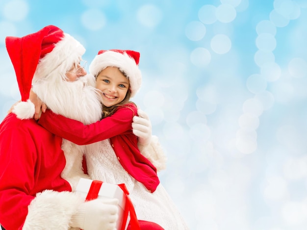 feriados, natal, infância e conceito de pessoas - menina sorridente abraçando com papai noel sobre fundo de luzes azuis