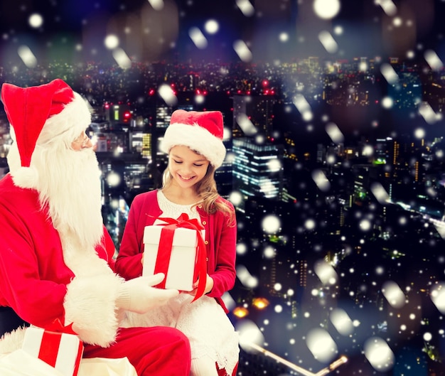 feriados, natal, infância e conceito de pessoas - menina sorridente com papai noel e presentes sobre o fundo da cidade nevada