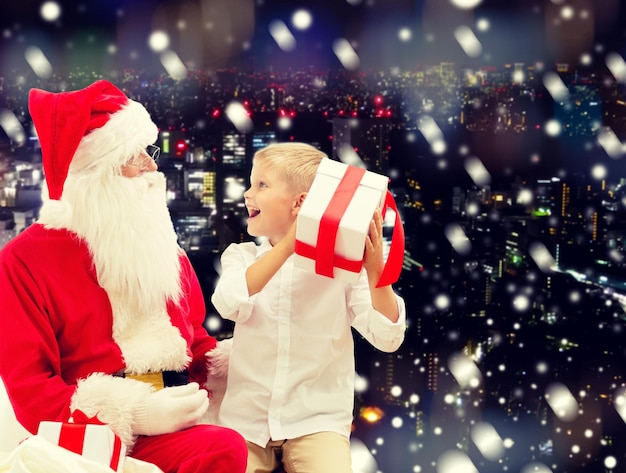 feriados, natal, infância e conceito de pessoas - menino sorridente com papai noel e presentes sobre o fundo da cidade nevada