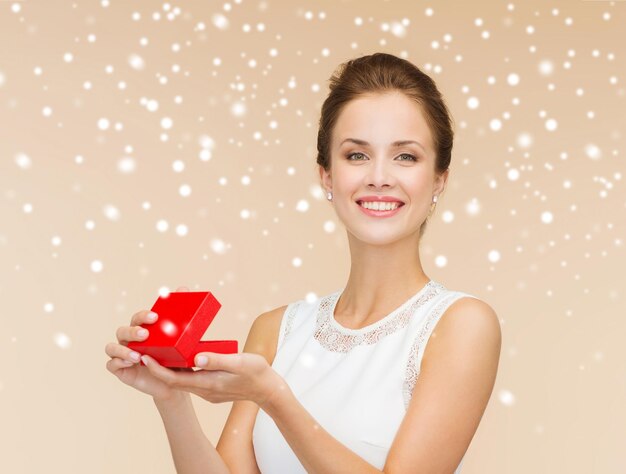 Feriados, presentes, casamento e conceito de felicidade - mulher sorridente de vestido branco segurando a caixa de presente vermelha sobre fundo bege sobre fundo bege e neve