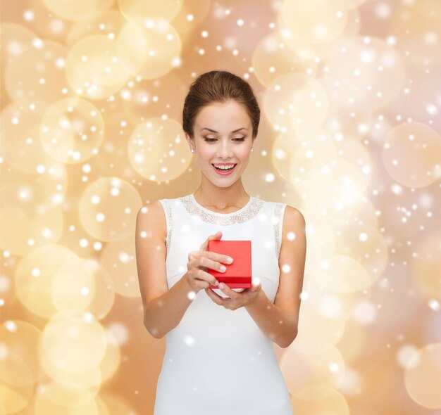 feriados, presentes, casamento e conceito de felicidade - mulher sorridente de vestido branco segurando a caixa de presente vermelha sobre fundo de luzes bege e neve