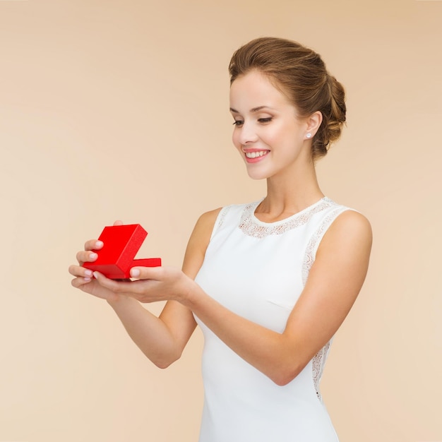 feriados, presentes, casamento e conceito de felicidade - mulher sorridente de vestido branco segurando caixa de presente vermelha sobre fundo bege