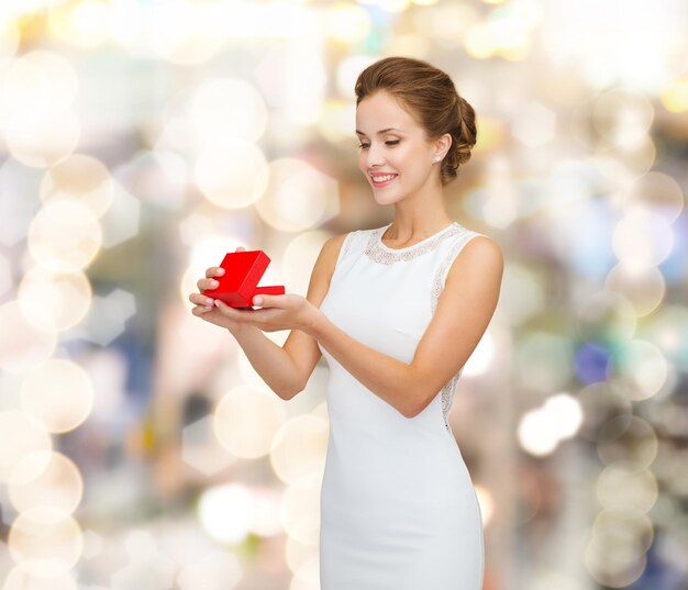 feriados, presentes, casamento e conceito de felicidade - mulher sorridente de vestido branco segurando caixa de presente vermelha sobre fundo de luzes brilhantes