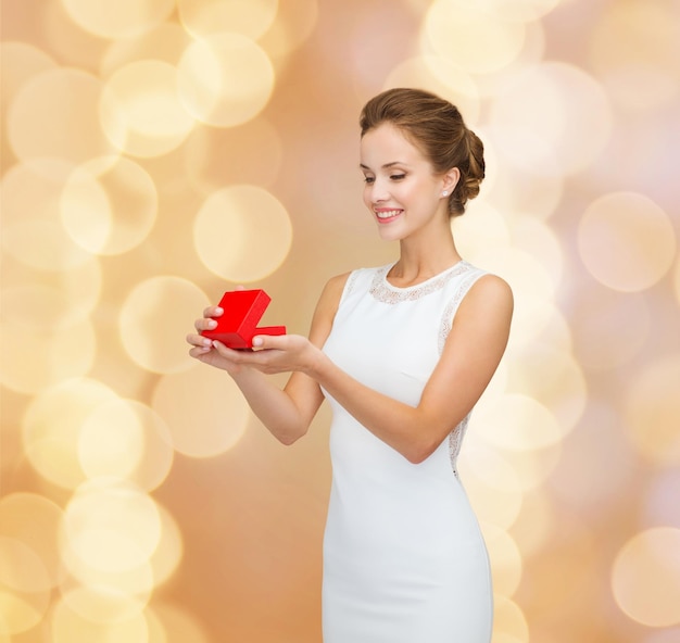 feriados, presentes, casamento e conceito de felicidade - mulher sorridente de vestido branco segurando caixa de presente vermelha sobre fundo de luzes douradas