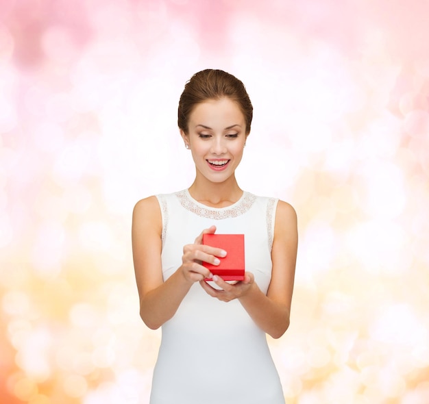 feriados, presentes, casamento e conceito de felicidade - mulher sorridente de vestido branco segurando caixa de presente vermelha sobre fundo de luzes rosa