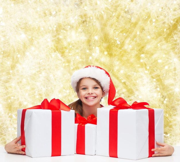 feriados, presentes, natal, infância e conceito de pessoas - garota sorridente no chapéu de ajudante de papai noel com caixas de presente sobre fundo de luzes amarelas