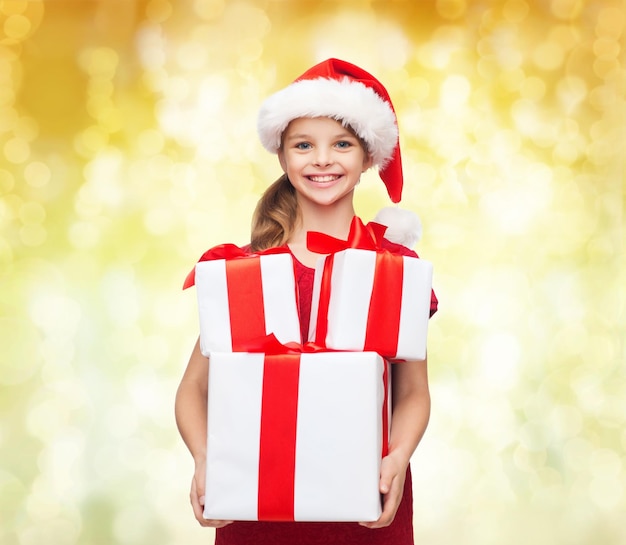 feriados, presentes, natal, infância e conceito de pessoas - menina sorridente no chapéu de ajudante de papai noel com caixas de presente sobre fundo de luzes amarelas