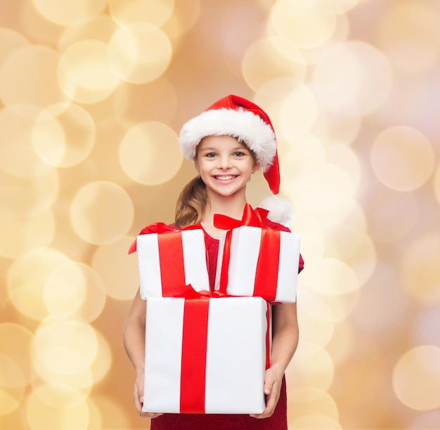 feriados, presentes, natal, infância e conceito de pessoas - menina sorridente no chapéu de ajudante de papai noel com caixas de presente sobre fundo de luzes bege