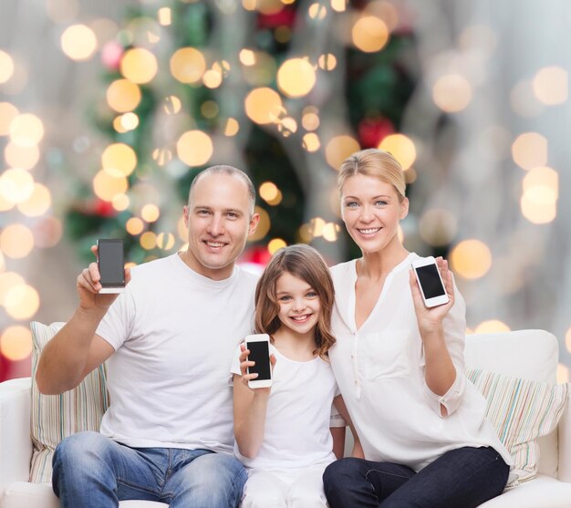 feriados, tecnologia, propaganda e conceito de pessoas - família sorridente com smartphones sobre a sala de estar e o fundo da árvore de natal