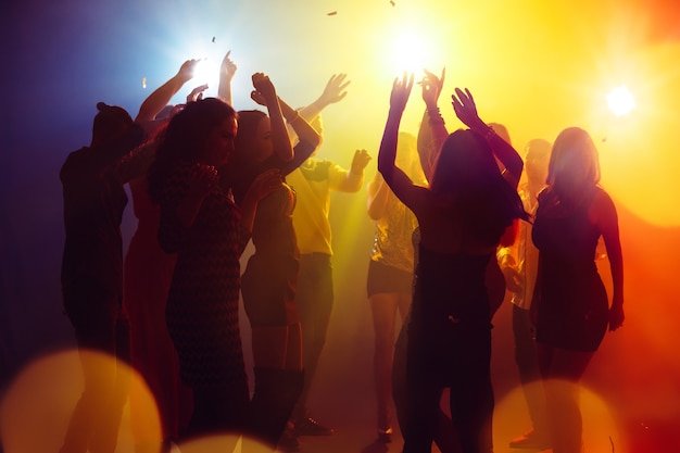Feriados. Uma multidão de pessoas em silhueta levanta as mãos na pista de dança em fundo de luz de néon. Vida noturna, clube, música, dança, movimento, juventude. Cores amarelo-azuladas e meninos e meninas em movimento.