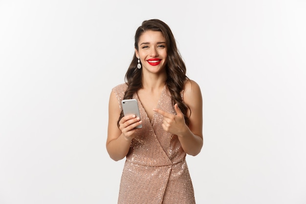 Férias, conceito de compra online. Mulher bonita satisfeita em vestido de luxo e batom vermelho, apontando para o celular, sorrindo satisfeita, fundo branco