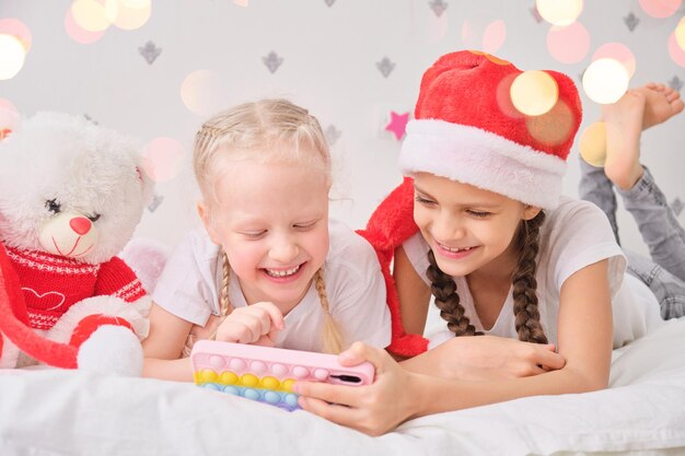 Férias de inverno e conceito de crianças - meninas felizes e sorridentes com smartphone deitado na cama. Luzes de natal