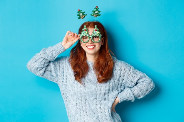 Férias de inverno e conceito de vendas de natal. modelo feminino ruiva linda comemorando o ano novo, usando óculos e bandana de festa engraçada, sorrindo para a câmera.