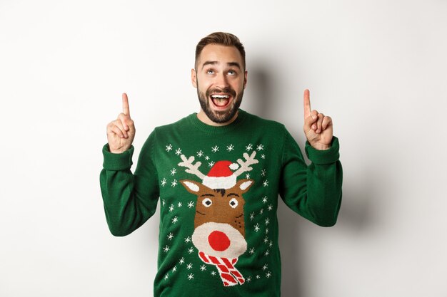 Férias de inverno e natal. homem barbudo animado em suéter engraçado olhando, apontando os dedos para o espaço da cópia, em pé sobre um fundo branco.