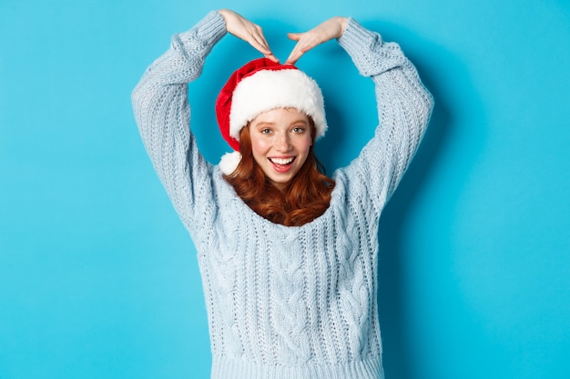 Férias de inverno e o conceito de véspera de Natal. Menina adolescente ruiva bonita com chapéu de Papai Noel e camisola, fazendo sinal de coração e sorrindo, desejando feliz Natal, em pé sobre fundo azul.