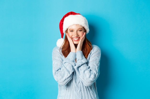 Férias de inverno e o conceito de véspera de Natal. Menina ruiva alegre com chapéu de Papai Noel, comemorando o ano novo, corando e sorrindo feliz, em pé sobre um fundo azul.