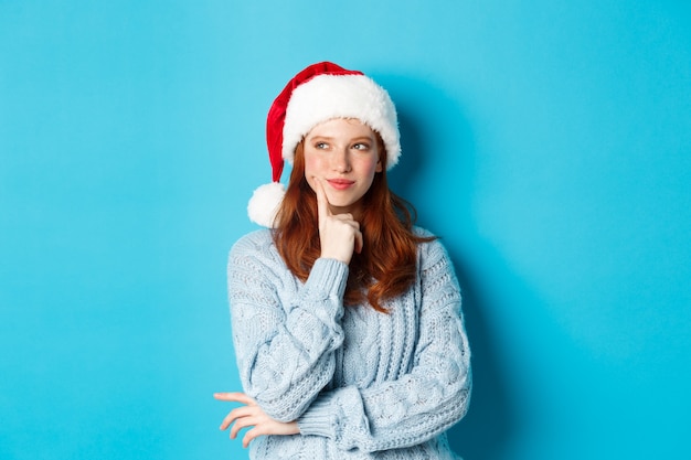 Férias de inverno e o conceito de véspera de Natal. Ruiva tola com sardas, usando chapéu de Papai Noel e pensando, planejando a celebração do ano novo, em pé sobre um fundo azul.