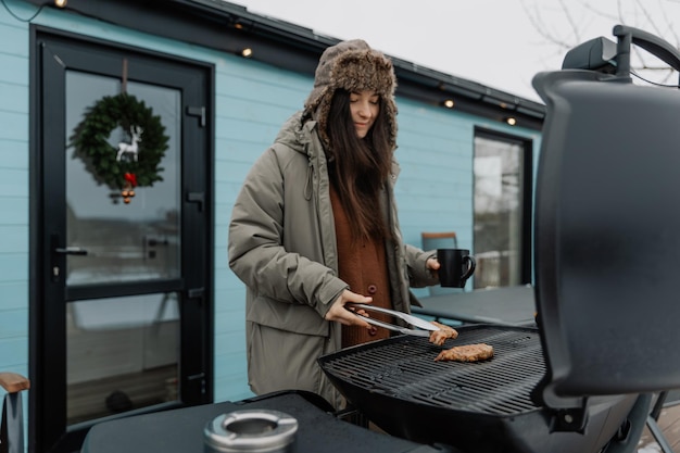 Férias de inverno no quintal de uma casa turística de campo Uma jovem mulher elegante está grelhando carne em uma grelha de gás para seus amigos e família