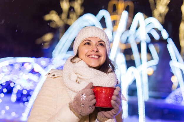 Férias de natal. Bela mulher sorridente em roupas quentes com uma xícara de café quente ao ar livre no inverno. Noite da cidade.