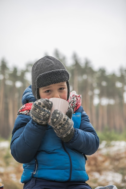 Férias de Natal, menino bebendo bebida quente de ano novo. Família feliz em uma caminhada ao ar livre na floresta ensolarada de inverno