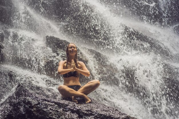 Férias de spa de bem-estar e conceito de meditação de ioga jovem mulher sentada em posição de lótus na rocha na cachoeira tropical