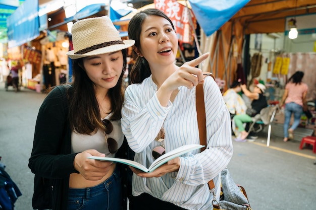 férias de verão city break e conceito de turismo. duas jovens coreanas asiáticas de chapéu de palha com amigos segurando o livro de guia de viagem e pesquisa de ponto para a famosa loja local para visitar no mercado tradicional