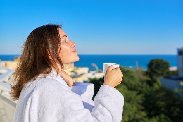 Férias de verão da mulher madura feliz Mulher de meia idade em roupão branco na varanda do hotel na cidade estância balnear Desfrutando de uma xícara de café sol bela paisagem marinha cópia espaço céu azul mar