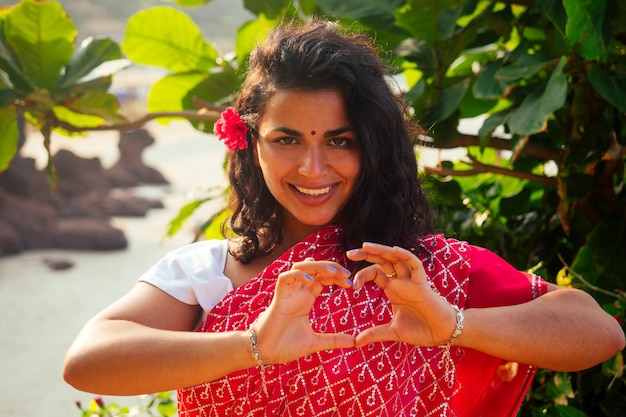 Férias de verão dia dos namorados na índia menina enviou beijo de ar e forma de coração linda mulher vestindo roupa tradicional indiana casamento vermelho saree sari em árvores tropicais paraíso ilha mar oceano praia