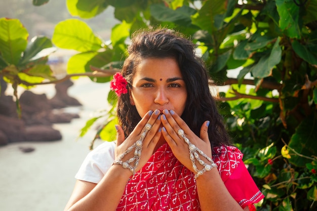 Férias de verão dia dos namorados na Índia menina enviou beijo de ar e forma de coração linda mulher vestindo roupa tradicional indiana casamento vermelho saree sari em árvores tropicais paraíso ilha mar oceano praia