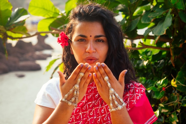 Férias de verão dia dos namorados na índia menina enviou beijo de ar e forma de coração linda mulher vestindo roupa tradicional indiana casamento vermelho saree sari em árvores tropicais paraíso ilha mar oceano praia