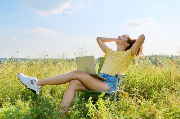 Férias de verão em mulher adulta de prado selvagem descansando na cadeira dobrável para acampar usando laptop