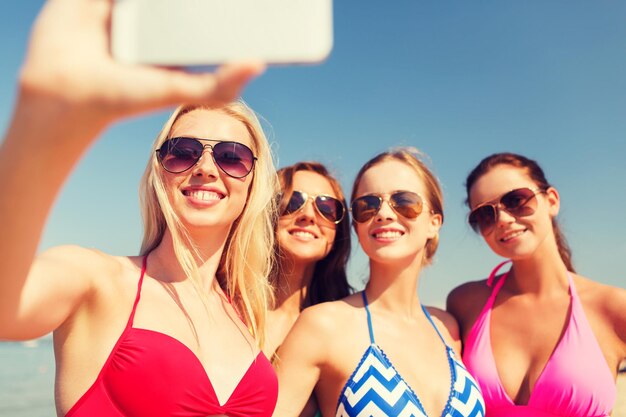 férias de verão, feriados, viagens, tecnologia e conceito de pessoas - grupo de mulheres jovens sorridentes na praia fazendo selfie com smartphone sobre fundo de céu azul