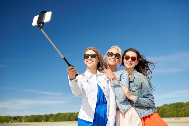 férias de verão, feriados, viagens, tecnologia e conceito de pessoas - grupo de mulheres jovens sorridentes tirando fotos com smartphone no bastão de selfie na praia