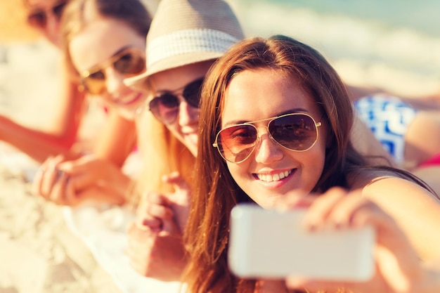 Férias de verão, viagens, tecnologia e conceito de pessoas - close-up de mulheres sorridentes em óculos de sol e chapéus fazendo selfie com smartphone na praia