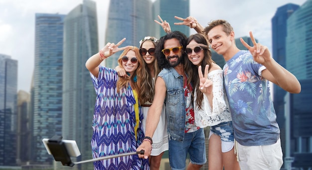 férias de verão, viagens, turismo, tecnologia e conceito de pessoas - jovens amigos hippies sorridentes tirando foto pelo smartphone selfie stick sobre o fundo da rua da cidade de singapura